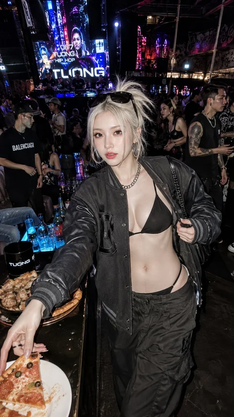 A blonde woman in black streetwear reaches toward a pizza among a crowd at a brightly lit nighttime venue.
